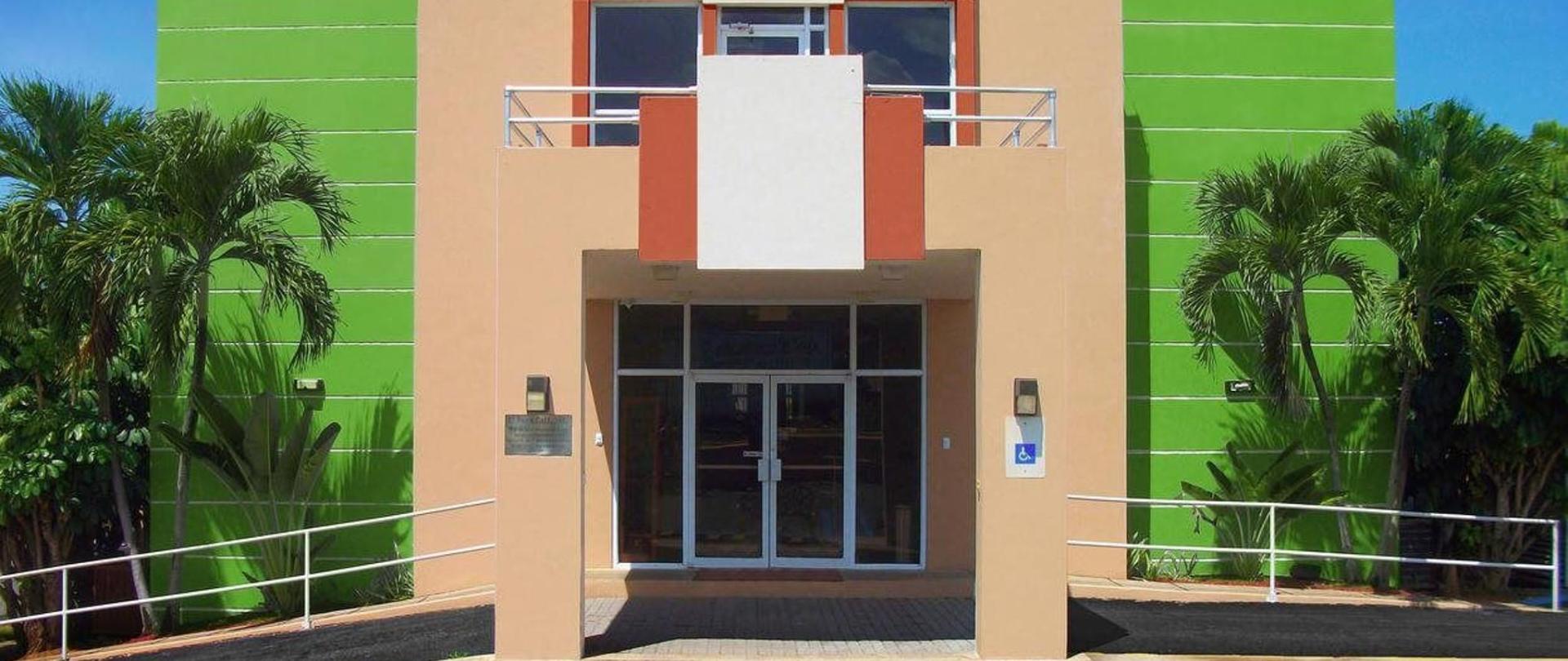 Coastal hotel entrance with striped beige and green exterior walls, orange accent panel, glass double doors framed by tropical palm trees under a bright sky