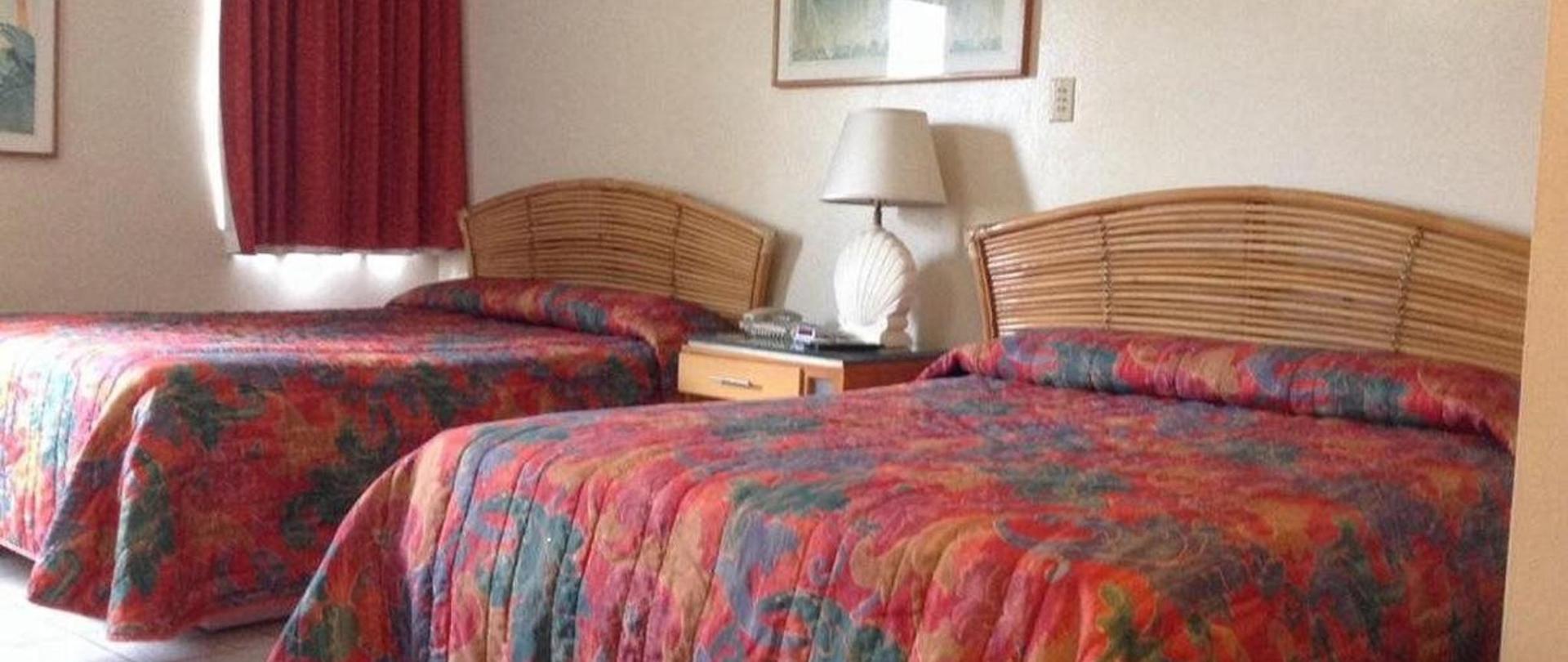 Two guest beds with floral patterned bedspreads and rattan headboards, a bedside lamp, and warm red curtains in soft amber light