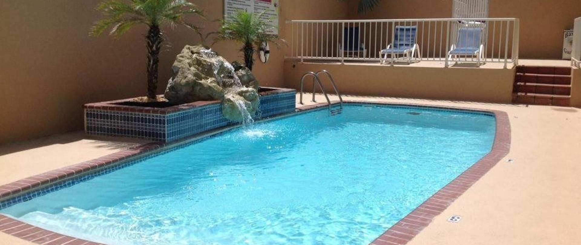 Outdoor swimming pool with a rock waterfall feature, tropical palms, and lounge chairs on an upper deck in blue and tan tones under a sunny sky