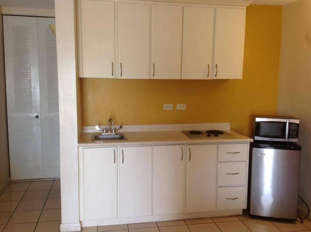 Small kitchenette with white cabinets, mustard yellow wall, sink, stovetop, microwave, and mini fridge on a tile floor