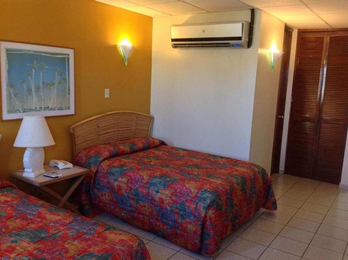 Hotel room corner with a floral bedspread, wall-mounted air conditioner, lamp, and wooden closet doors in warm tones