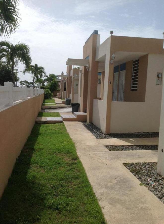 Exterior walkway along a row of small stucco buildings with palm trees, a white perimeter wall, and green lawn under a bright sky