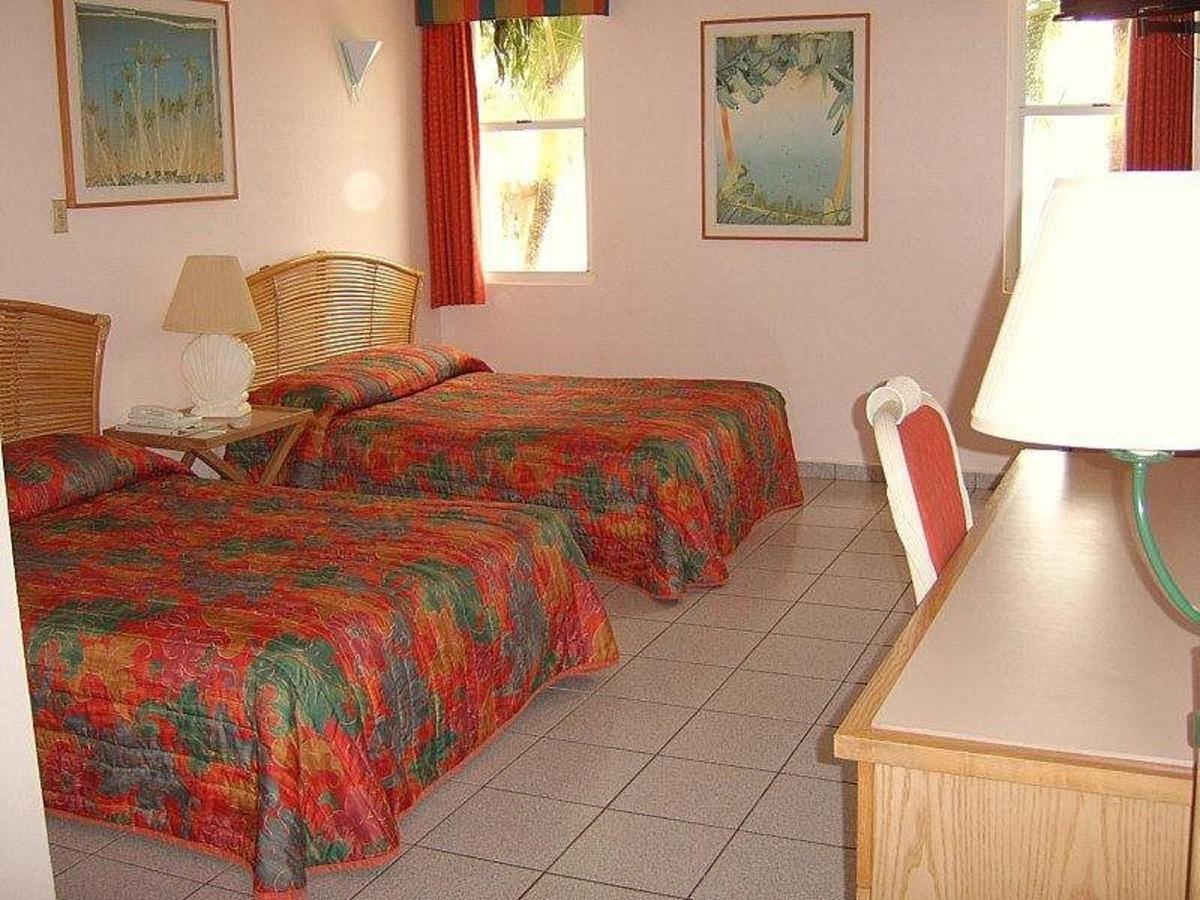 Hotel room with two beds featuring colorful floral bedspreads, tan tile floor, pink walls, and framed tropical artwork