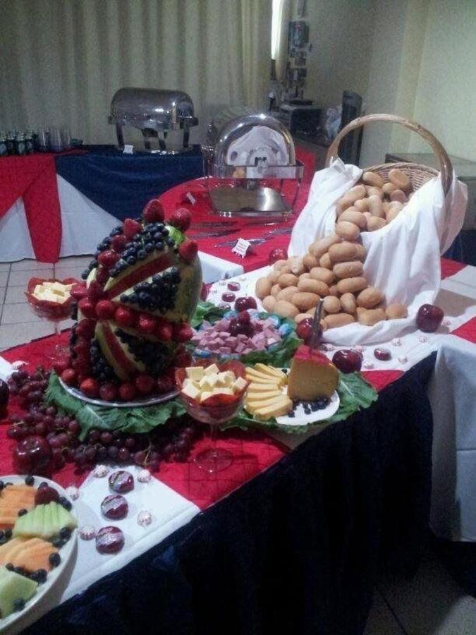 Buffet spread with a carved fruit centerpiece, cheese and meat platters, a bread basket, and red and dark blue table linens